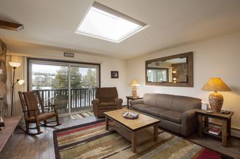 The living area of Hi Country Haus 2115 features a comfortable sofa, recliner, rocking chair, coffee table, and a large mirror, with a balcony offering snowy outdoor views.