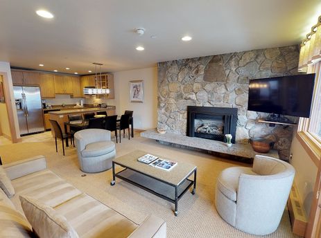 A spacious living room featuring a stone fireplace, cozy seating, and an adjacent kitchen in the Manor Vail Lodge Platinum 3 Bedroom Condo.