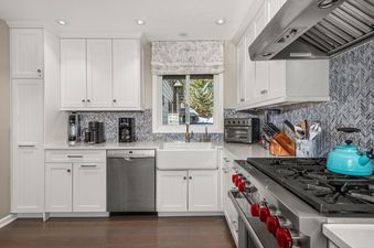 A modern kitchen with white cabinetry, a chevron backsplash, stainless steel appliances, and a farmhouse sink under a window with a snowy view.