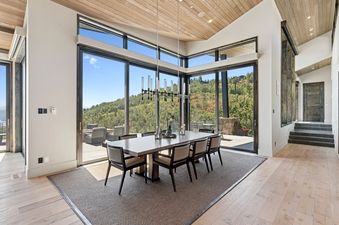 A modern dining room in Star Gazer at Deer Valley features a large table with eight chairs, illuminated by a linear chandelier, and surrounded by floor-to-ceiling windows offering expansive views of green hills and blue sky.