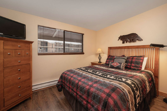 A cozy bedroom in Beaver Village 333 featuring a comfortable bed with plaid bedding, a wooden dresser with a television, and a window with blinds.