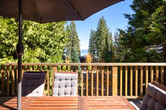 A private balcony with a wooden table and two chairs under a large umbrella, overlooking a scenic view of pine trees and a distant lake.