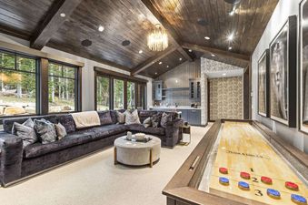 The games room at Ruby Run features a large sectional sofa, a shuffleboard table, a wet bar, and expansive windows showcasing the scenic outdoors.