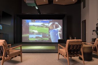 An indoor golf simulator room at Over the Edge, featuring a large screen displaying a golf course and golfer, surrounded by comfortable leather armchairs.
