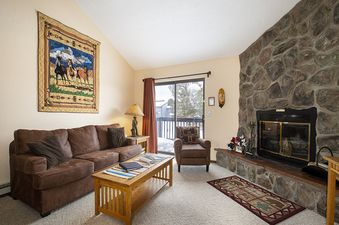 The living area of Meadow Ridge 28 Unit 6 features a comfortable brown sofa, a wooden coffee table, a grand stone fireplace, and a large window with an outdoor view.