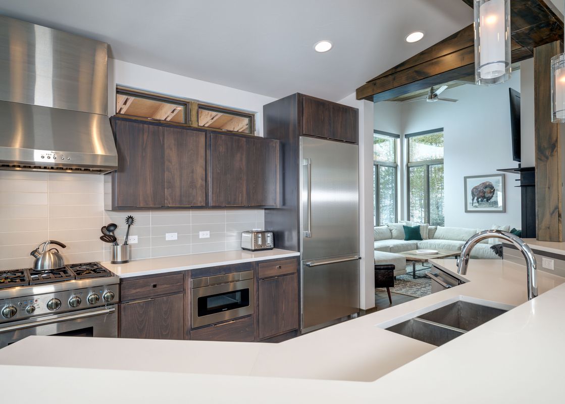 A modern kitchen with dark wood cabinets, white countertops, and stainless steel appliances, featuring a large refrigerator, gas range, and a dual-basin sink.