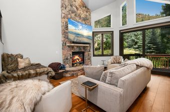 A cozy living room in Chateau Tremonte 8 with a stone fireplace, television, and plush seating, offering views of pine trees and mountains through large windows.