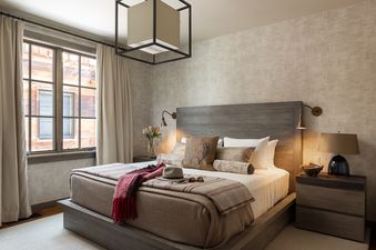 A cozy bedroom with a large bed, wooden headboard, stylish bedding, bedside tables with lamps, and large windows with drapes.