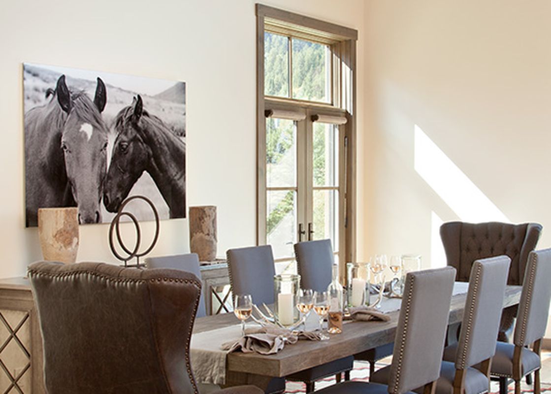 A spacious dining area with a long wooden table, leather and upholstered chairs, a patterned rug, and a horse art print on the wall, with natural light entering from tall windows.