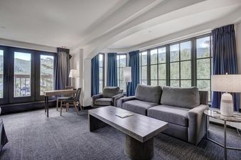 The Pines Lodge King Corner Room showcases a luxurious living space with a large grey sofa, armchairs, a wooden coffee table, and a dining area, all set against a backdrop of scenic mountain views through floor-to-ceiling windows.