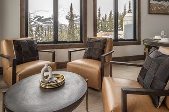 A cozy living area at 22 Lower Cascade Ridge features three leather armchairs, a coffee table, and large windows with views of snow-covered mountains and evergreen trees.