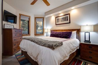 A king bedroom at Chateau Chamonix CX322 with a large wooden bed, white bedding, purple and grey accents, a wooden dresser with a TV, and two windows.
