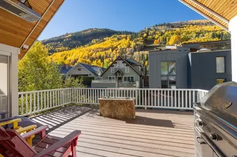 A spacious outdoor deck with colorful Adirondack chairs, a BBQ grill, and a stone fire pit, offering stunning views of a mountain covered in yellow autumn trees.
