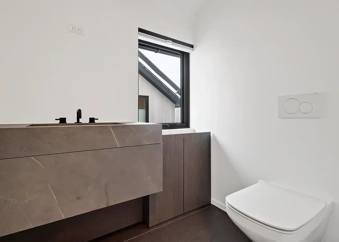 Minimalist bathroom in Maison Cornet featuring a gray stone vanity, black faucet, wall-hung toilet, and a window showing the exterior roofline.