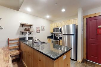 A modern kitchen with light wood cabinets, stainless steel appliances, and a granite-topped island with a sink, all well-lit by recessed ceiling lights.