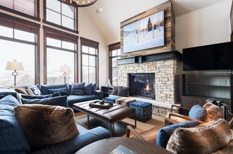 A spacious and elegantly furnished living room in Silver Star 40, featuring a stone fireplace, comfortable blue seating, and large windows with a winter view.