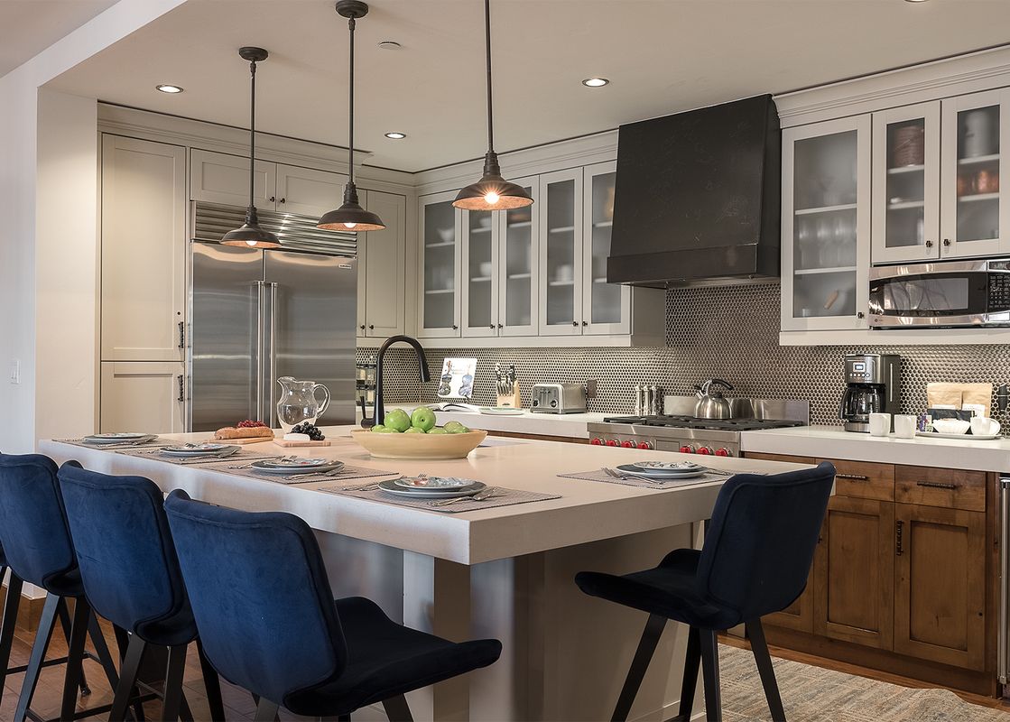 A modern kitchen with a large island, blue velvet bar stools, stainless steel appliances, and elegant pendant lighting.
