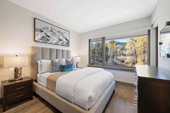 The bedroom of Riverfront Residences 02 features a large bed with a fabric headboard, two nightstands with lamps, a dresser, and a large window offering an autumn view.