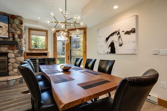 A modern dining room with a large wooden table, black leather chairs, and a contemporary chandelier, complemented by a stone fireplace and a framed artwork of a horse.