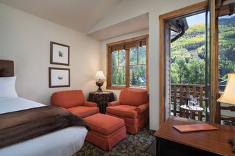 A cozy bedroom with a bed, two orange armchairs, and a window and balcony overlooking a scenic mountain view.