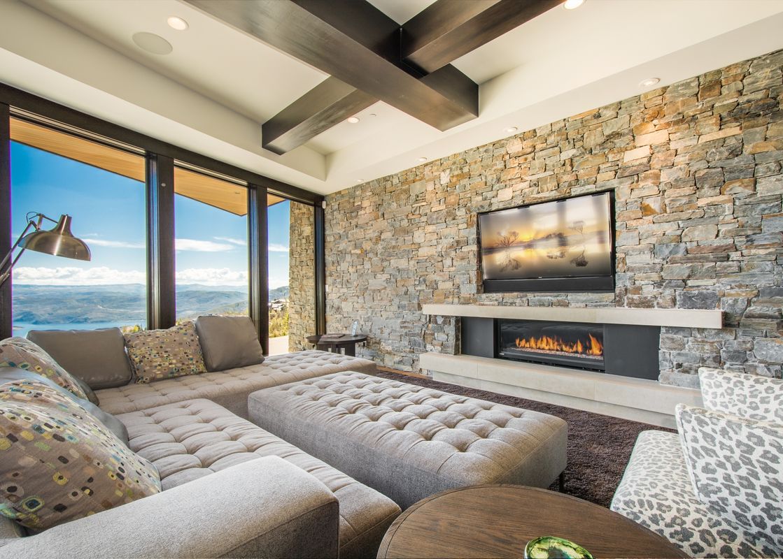 A spacious living room at Deer Crest Estate with a stone feature wall, fireplace, large sectional sofa, and expansive windows offering a stunning mountain and lake view.