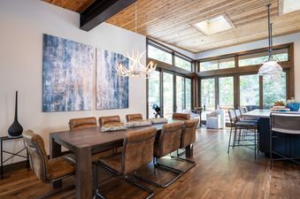 A dining area with a long wooden table, brown leather chairs, and an antler chandelier, with large windows overlooking a forest.
