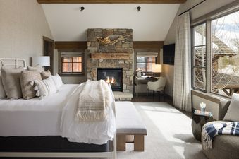 A master bedroom in Fish Creek Lodge 05 features a large bed, a stone fireplace, a small desk area, and windows overlooking an outdoor scene.