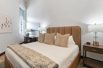 A tastefully decorated bedroom in Powder Run B32 featuring a large bed with a brown headboard, white linens, neutral-toned pillows, and a patterned throw blanket, alongside a nightstand with a lamp and an adjacent desk area with a chair and artistic decor