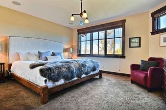 The primary bedroom at Silver Star 102 features a large bed with a white headboard, dark furry throw, and expansive windows, alongside a red armchair and dark wood dresser.