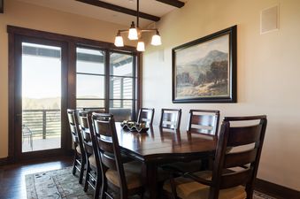 A dining room at Silver Star 1302 with a large wooden table and chairs, a scenic painting on the wall, and glass doors leading to an outdoor balcony with mountain views.