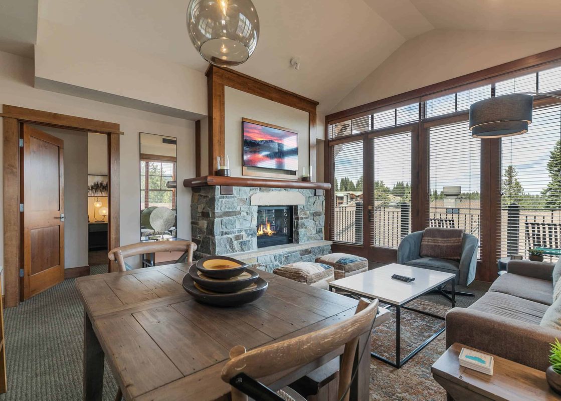 A spacious living room with a fireplace, large windows overlooking a wooded area, and a dining table in the foreground.