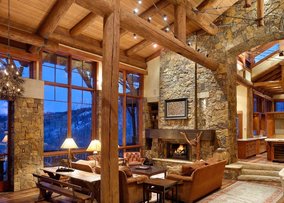 A rustic great room with a stone fireplace, large windows, and timber beams, offering mountain views.