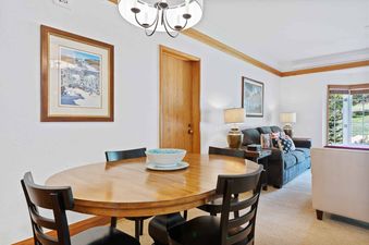 A cozy dining area with a wooden oval table and black chairs, transitioning into a living room with a gray sofa, patriotic pillows, and a large window looking out onto green foliage at The Charter B2305.