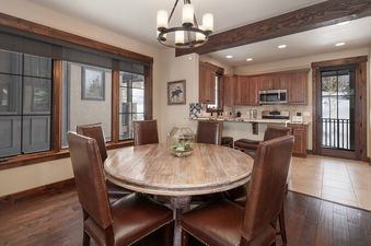 A rustic-chic dining area with a round wooden table surrounded by six brown leather chairs, featuring a kitchen in the background.