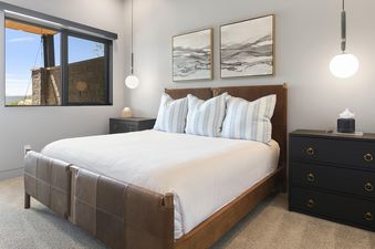 A luxurious bedroom in SkyRidge 1868, featuring a king-sized bed with white linens and striped pillows, flanked by dark wooden nightstands and modern pendant lights, all set against a backdrop of light gray walls and a large window offering an exterior vi