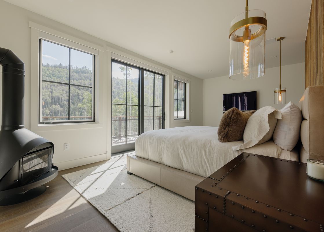 A bright and airy master bedroom with a king-size bed, a modern black fireplace, and large windows offering a view of a forested mountain.