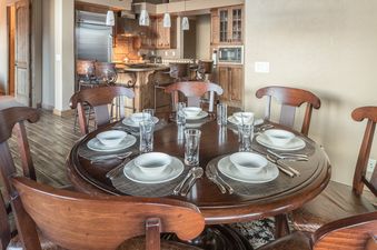 A round wooden dining table is set for a meal in a spacious dining area that opens up to a modern kitchen.