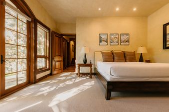 A luxurious bedroom at Four Bears Hideaway at See Forever, featuring a large bed, wooden furniture, and sunlit windows opening to a stone wall exterior.