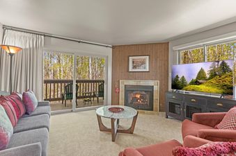 The inviting living room at Snow Flower 140 Park City features a cozy fireplace, comfortable seating, and a large flat-screen TV, all opening onto a balcony with a scenic wooded view.