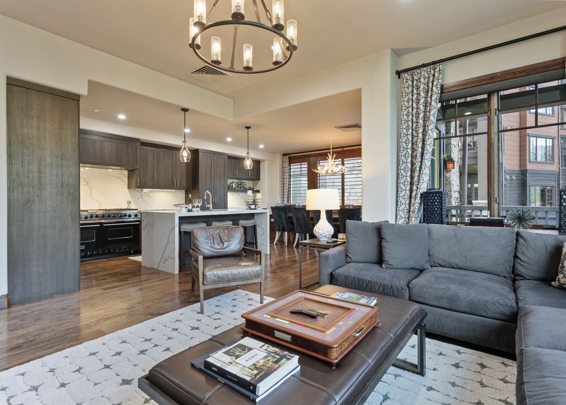 A spacious open-concept living area with a grey sectional sofa, brown leather ottoman, and a modern kitchen with dark wood cabinets and marble countertops.