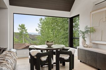 A modern dining area at Onyx Lodge with a round table, dark green chairs with white faux fur, and large corner windows offering expansive views of green mountains.