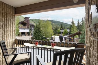 A close-up view of a balcony, with two chairs and a unique snowboard-shaped table holding two red mugs, overlooking a charming village with buildings, trees, and mountains in the background.