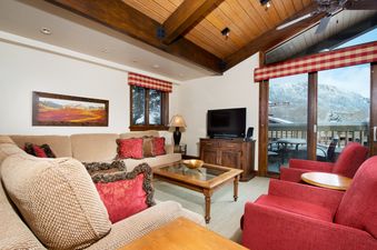 The living area of The Gant Aspen 4 Bedroom Premier features a comfortable L-shaped sofa and red armchairs, with a large glass door opening to a balcony overlooking snow-covered mountains.