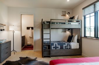 A bunk room with a wooden bunk bed, a dresser, a floor lamp, and a window with an outdoor view.