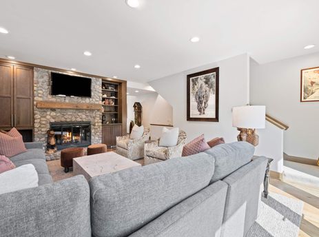 A cozy living room in Village Hall 13 features a stone fireplace, comfortable sofas, and rustic decor, illuminated by recessed lighting.