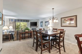 An inviting open-concept living and dining area in Snow Flower 135 Park City features a dining table with six chairs, a cozy living room with patterned sofas, a fireplace with a TV, and a large window overlooking lush trees.