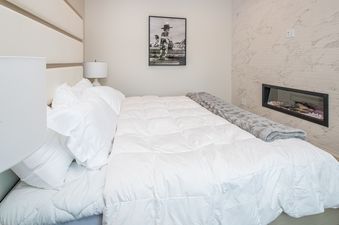 A serene bedroom at 7 Lower Cascade Ridge with a plush white bed, a gray throw, bedside lamps, a framed black and white artwork, and a modern fireplace.