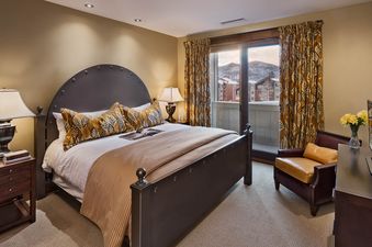 The bedroom at Emerald Mountain One Steamboat Place 702 features a large bed with a dark metal frame, patterned pillows, a patterned throw, and a nightstand with a lamp. A sliding glass door leads to a balcony with mountain views.