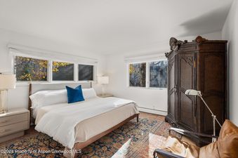 A bedroom with a large bed, a chair, an armoire, and two windows with fall foliage views.