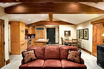 An open concept living space featuring a comfortable sofa, a fully equipped kitchen with wooden cabinets, and a dining area with a round table and chairs, all under rustic wooden beams.